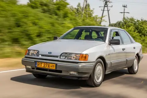 Srebrny Ford Scorpio jedzie po drodze, otoczony zielenią.