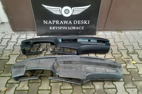Dwa kokpity samochodowe, jeden czarny, drugi szary, leżą na chodniku przed tablicą z napisem "NAPRAWA DESKI".