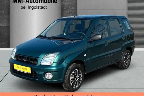 Zielony Subaru Justy z żółtą tabliczką "TOP ZUSTAND" na tle hali dealerskiej.