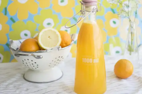 Świeżo wyciśnięty sok pomarańczowy w butelce, obok pomarańczy i cytryny w sitku. Jak dbać o świeżość owoców, by uniknąć objawów sfermentowanego soku w barze.