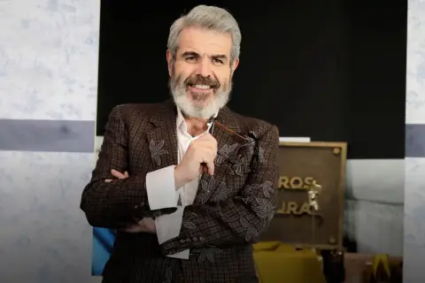 Lorenzo Caprile, con su característica barba canosa, sonríe mientras sostiene unas gafas. Viste una chaqueta de tweed con bordados florales.