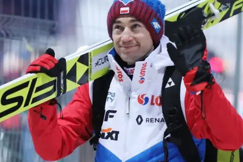Uśmiechnięty Andrzej Stoch Jr. z nartami na ramieniu, gotowy do skoku.