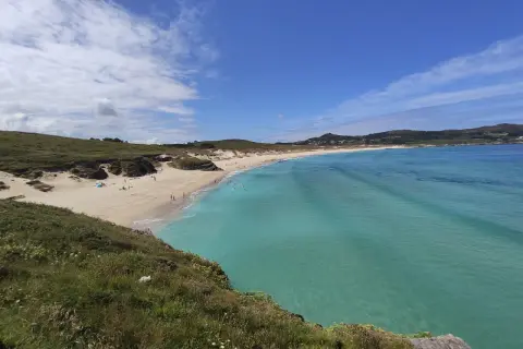 Vista panorámica de la playa de Santa Comba, con arena dorada, aguas turquesas y colinas verdes. Algunas personas disfrutan del sol y el mar.