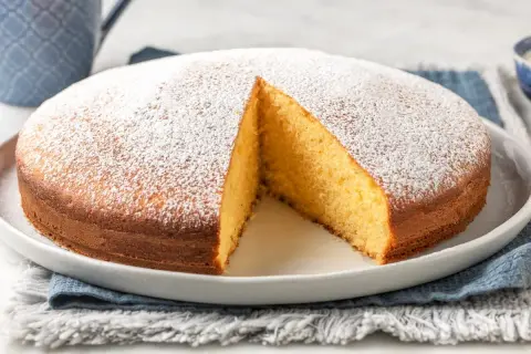 Torta yogurt greco soffice e dorata, cosparsa di zucchero a velo. Una fetta è stata tolta, rivelando l'interno profumato.