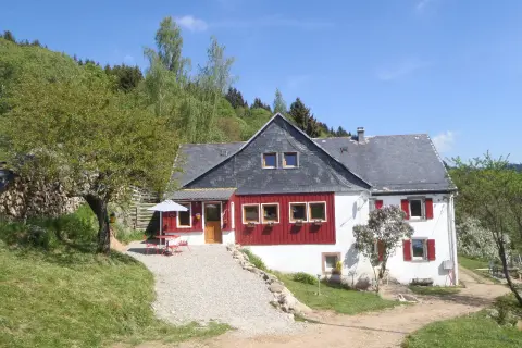 Maison de campagne avec terrasse et parasol, idéale pour un camping à la ferme dans les Vosges. Le soleil brille sur ce paysage verdoyant.