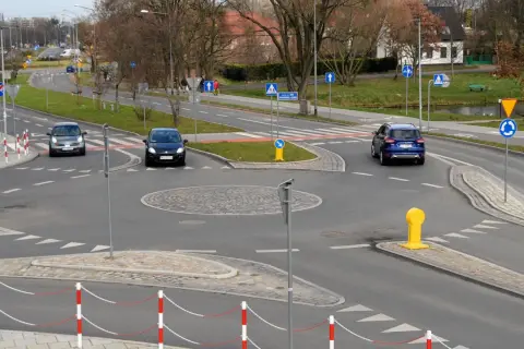 Samochody zbliżają się do ronda. Widoczny jest kierunkowskaz na rondzie, a także znaki drogowe i bariery.
