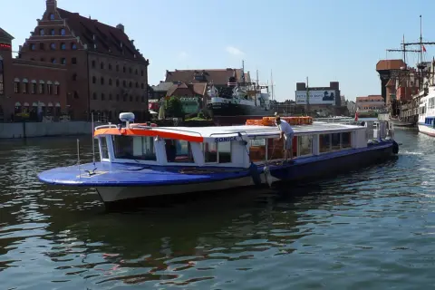 Niebiesko-biały tramwaj wodny "Sonica" płynie po Motławie w Gdańsku, mijając zabytkowe kamienice i inne statki.