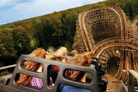 Fahrt mit einer Holzachterbahn, Menschen mit wehenden Haaren, im Hintergrund Wald und Himmel.