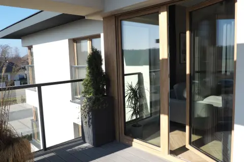 Nowoczesne okna dzielone z drewnianymi ramami otwierają widok na balkon z roślinnością i wnętrze domu.