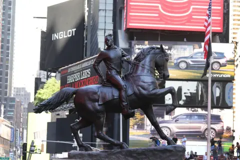 A bronze statue of a person on horseback stands in Times Square, with billboards for Inglot and Harper Lee's "To Kill a Mockingbird" in the background.