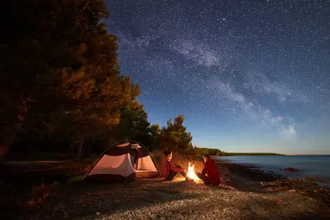 Nocne biwakowanie pod rozgwieżdżonym niebem. Dwoje ludzi siedzi przy ognisku obok namiotu z materiału namiotowego, na kamienistej plaży.