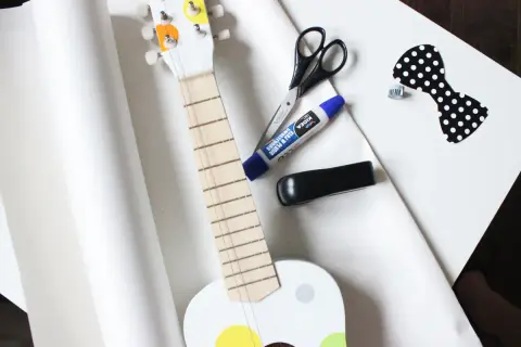 Przygotowania do wysyłki gitary: biała gitara, nożyczki, klej, zszywacz i ozdobna kokardka.