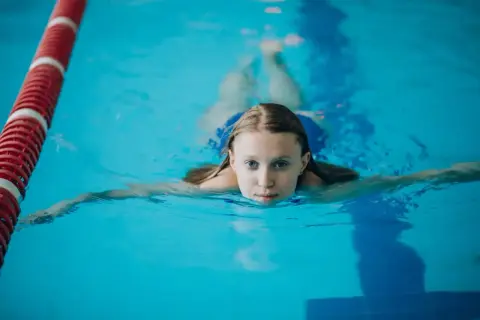 Dziewczynka ćwiczy pływanie żabką w basenie. Woda jest błękitna, a obok niej biegnie czerwono-biała lina.