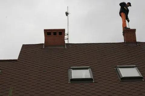 Mężczyzna w pomarańczowych spodniach stoi na kominie i czyści go szczotką.