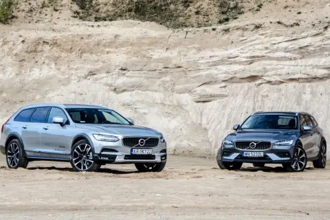Dwa samochody Volvo Cross Country, srebrny i grafitowy, stoją na piaszczystym podłożu przed piaskową ścianą.