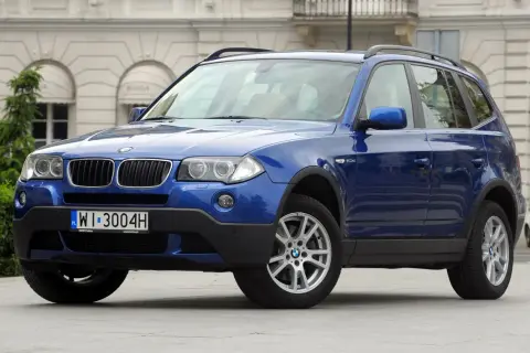 Niebieskie BMW X3 2005 z polskimi tablicami rejestracyjnymi WI 3004H zaparkowane przed budynkiem.