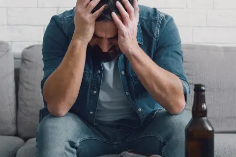 Man met handen in het haar, zittend op een bank met een fles bier. Dit illustreert de definitie van verslaving: een dwangmatige behoefte.