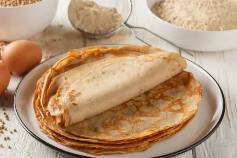 Stos naleśników gryczanych na talerzu, obok jajek i mąki.