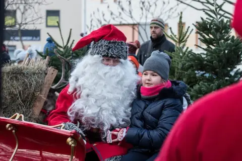 Święty Mikołaj w saniach z dziewczynką, która z zaciekawieniem patrzy przed siebie. W tle widać renifera i choinki.