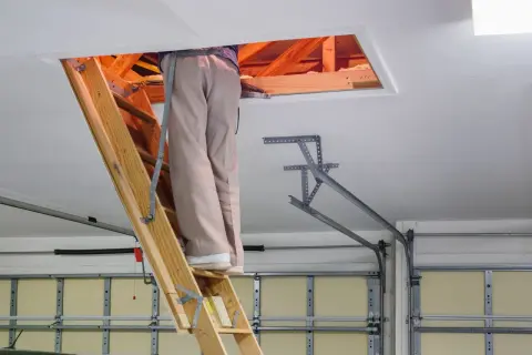 Osoba na drabinie przy wyłazie strychowym. Dowiedz się, jak uszczelnić wyłaz strychowy, by oszczędzać energię.