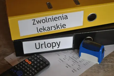 Zółty segregator "Zwolnienia lekarskie" i czarny "Urlopy" obok kalkulatora i pieczątki. Dokumenty na biurku sugerują, że kod a na zwolnieniu jest w trakcie rozliczeń.