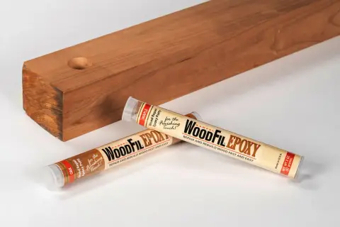 Two tubes of WoodFil Epoxy, a wood repair putty, lie next to a wooden plank with a hole, demonstrating how to use wood filler for quick repairs.