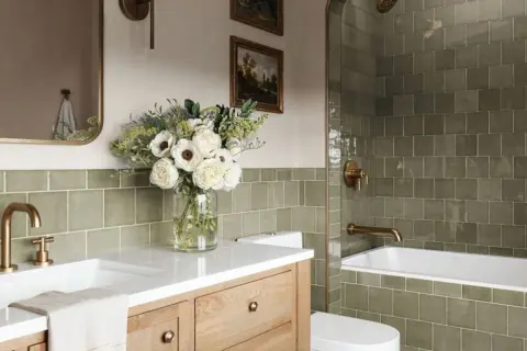 Élégante salle de bain verte avec carrelage texturé, vasque en bois, bouquet de fleurs blanches et robinetterie dorée.