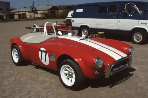 Röd sportbil, en kungens AC Cobra med nummer 77, redo för race. Bakom skymtar en blåvit skåpbil.