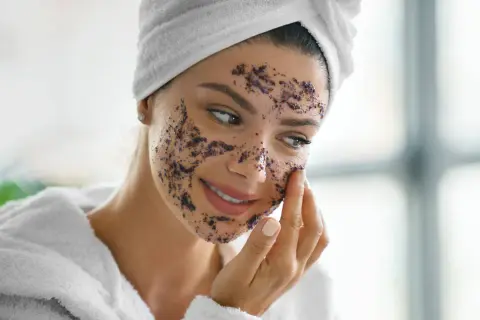 Femme souriante appliquant un gommage sur son visage. Elle porte une serviette sur la tête et un peignoir. La question est : combien de temps laisser un gommage sur le corps ?