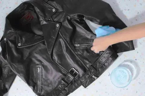 Schwarze Lederjacke wird mit einem feuchten Tuch gereinigt. Ein Schälchen mit Seifenwasser steht daneben. Lederjacke waschen.