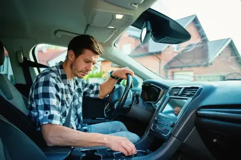 Mężczyzna w kratę siedzi za kierownicą, zastanawiając się, jak ruszyć autem. Jego ręka spoczywa na dźwigni zmiany biegów.