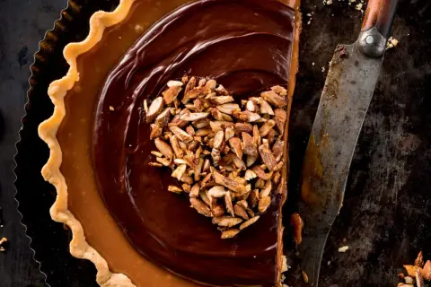 Herzförmige Schoko Tarte mit Karamell und gehackten Mandeln, daneben ein altes Messer.