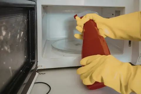 Manos con guantes amarillos rociando el interior de un microondas sucio con un limpiador.