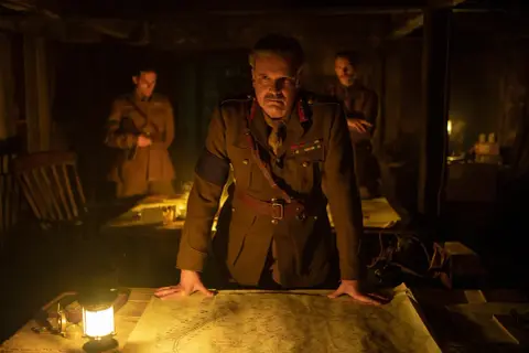 Ein General studiert im Schein einer Lampe eine Karte. Die besetzung von 1917 (film) zeigt ihn mit zwei Offizieren im Hintergrund.