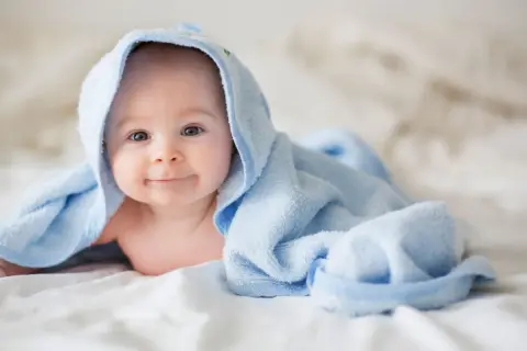 Ein lächelndes neugeborenes Baby liegt auf einem weißen Bett, eingewickelt in ein hellblaues Handtuch.