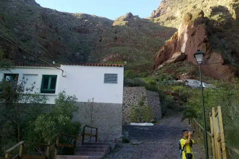Un niño toma fotos en un camino empedrado, con una casa blanca y montañas rojas de fondo, cerca de la cueva bermeja.