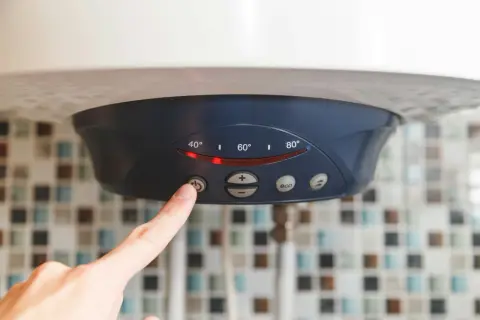 Palec naciska przycisk na bojlerze, zastanawiając się, czy bojler elektryczny pobiera dużo prądu. Wyświetlacz pokazuje temperaturę.