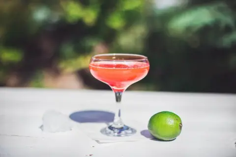 Orzeźwiający strawberry daiquiri w kieliszku, obok limonka i lód, na tle zieleni.