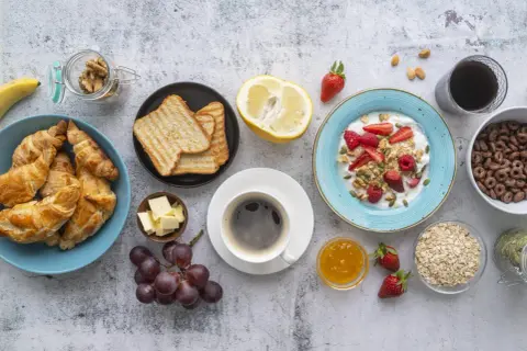 Un tavolo pieno di idee su cosa mangiare a colazione: croissant, toast, yogurt con frutta, caffè, uva e cereali.