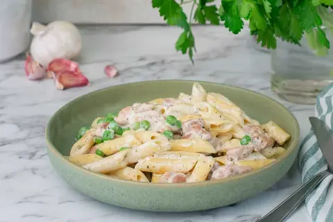 En krämig kyckling pasta med ärtor i en grön skål, med vitlök och persilja i bakgrunden.