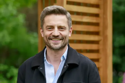 Uśmiechnięty Marcin Rogacewicz, znany z wielu filmów, pozuje na tle drewnianej pergoli i zieleni.