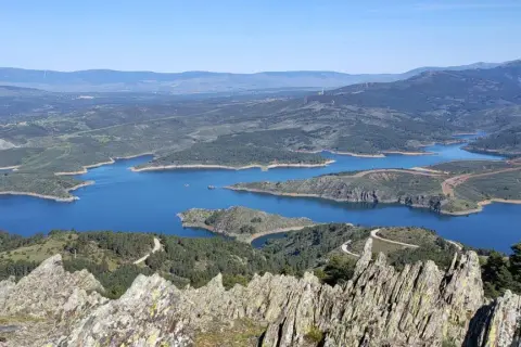 Vista panorámica del embalse de Puentes Viejas, con sus aguas azules serpenteando entre colinas verdes y montañas al fondo.