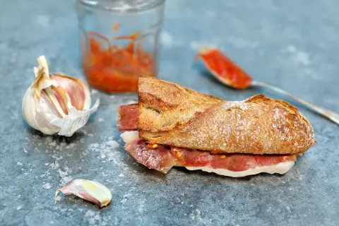 A delicious bocadillo de jamon, with a crusty baguette filled with cured ham, sits on a slate surface next to garlic and a spoonful of tomato spread.