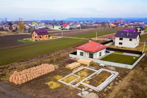 Budowa domku na działkę zrób to sam. Widać fundamenty, sterty cegieł i drewno budowlane. W tle ukończone domy.