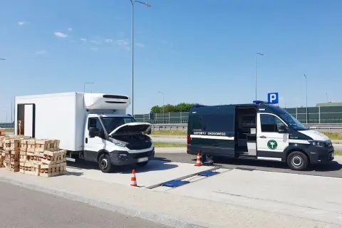 Kontrola drogowa: biała ciężarówka z otwartą maską i stosy palet obok, obok zielony bus Inspekcji Drogowej. Sprawdzana jest ładowność w dowodzie rejestracyjnym.