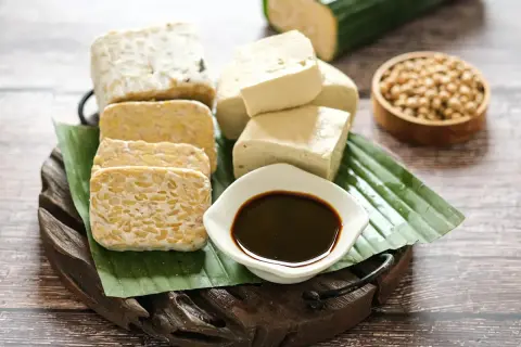 Tempeh und Tofu, zubereitet aus indonesischen Sojabohnen, liegen auf Bananenblättern. Dazu eine Schale dunkler Sojasauce und rohe Sojabohnen.