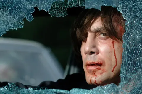 Javier Bardem w jednym z mrocznych filmów, z krwawiącą twarzą, patrzy przez pękniętą szybę.