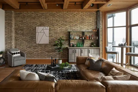 A rustic modern living room with exposed brick, wood beams, a leather sofa, and a home bar.