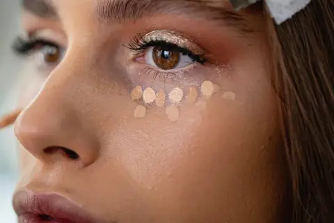 Nahaufnahme einer Frau, die Concealer unter den Augen aufträgt. Mehrere helle Tupfer bilden eine Reihe, bereit zum Verblenden.