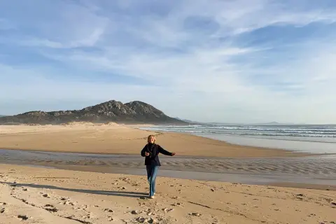 Mujer corre en la playa de Muros, Galicia, con montañas al fondo y el mar azul.